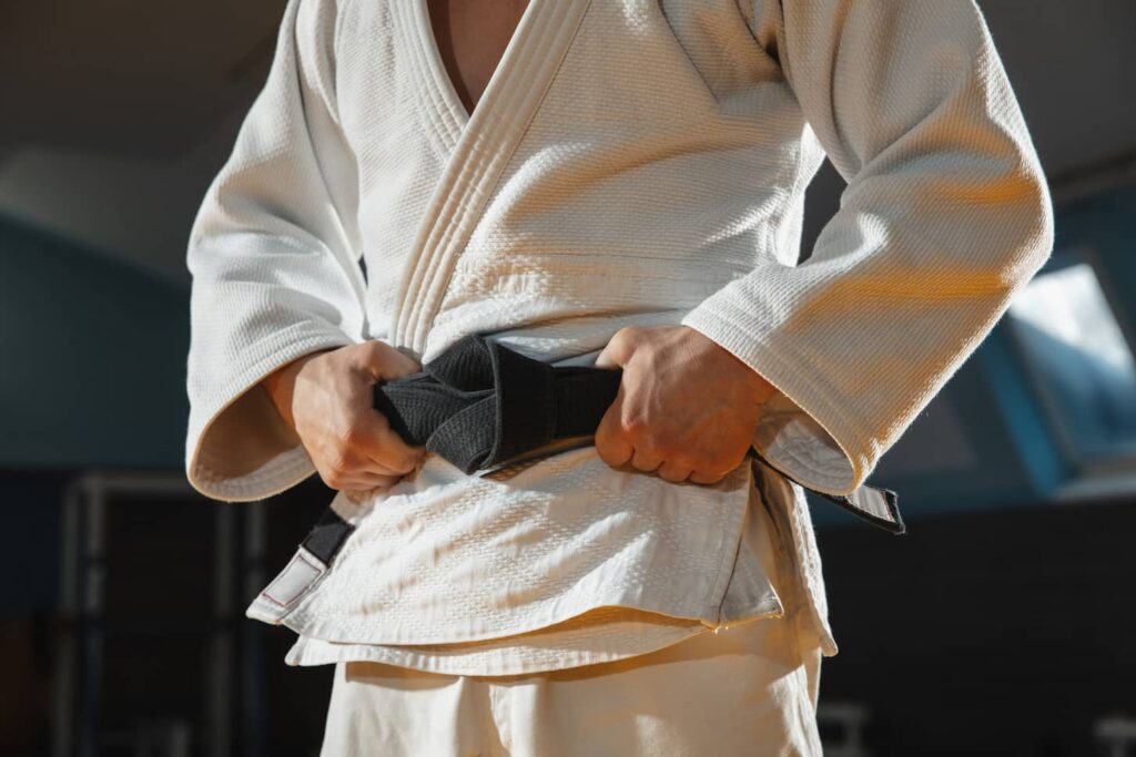 An image of a Young judo caucasian fighter in blue kimono with black belt posing confident in the gym, strong and healthy. Practicing martial arts fighting skills. Overcoming, reaching target, self building up.