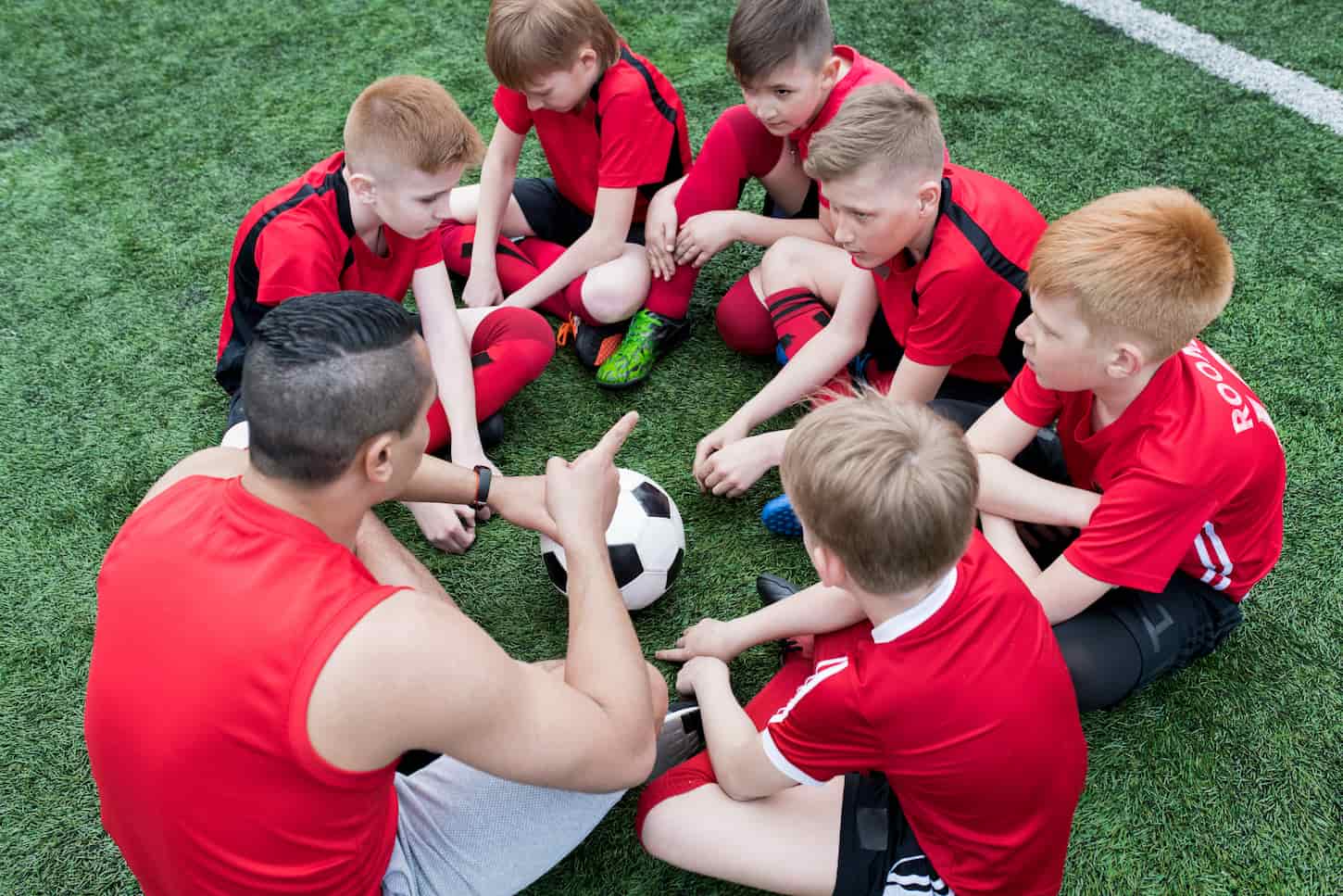 An image of a group of boys sitting in a circle listening to a motivational pep talk from the coach before team practice.