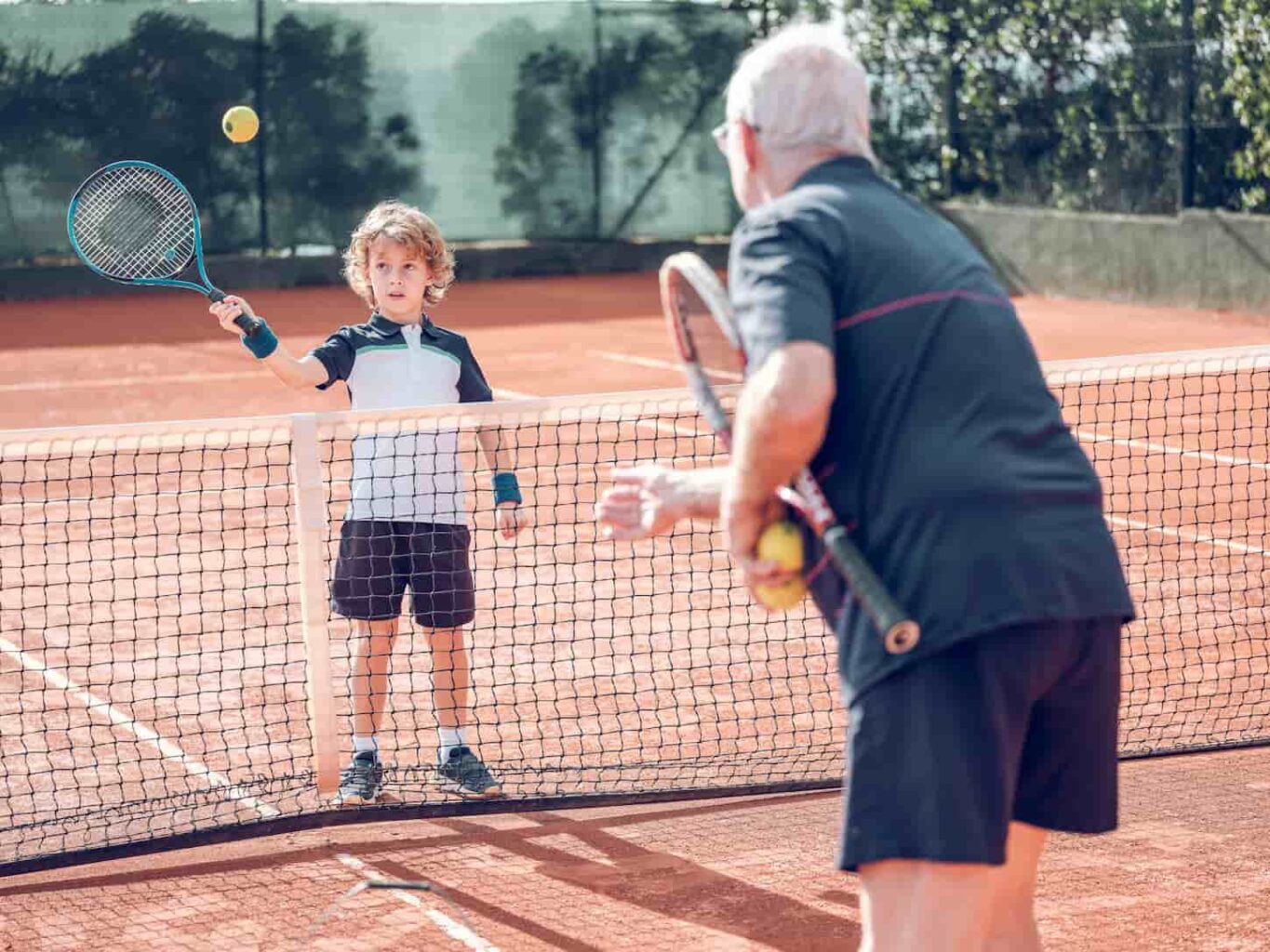 An image of a senior male training in sports clothes explaining how to hit the ball with a racket to the attentive little boy during tennis class on an outdoor court.