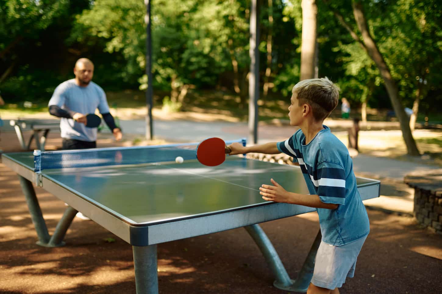 An image of a Father and son playing table tennis outdoors. The family leading a healthy lifestyle, daddy and boy, ping pong training in summer park.