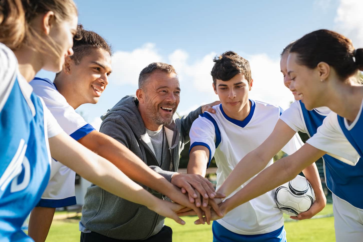 An image of a Happy coach and high school soccer team with their hands stacked on the football court. Young football players stacking hands together. A mature man coach encourages his students to do their best during the sport match.