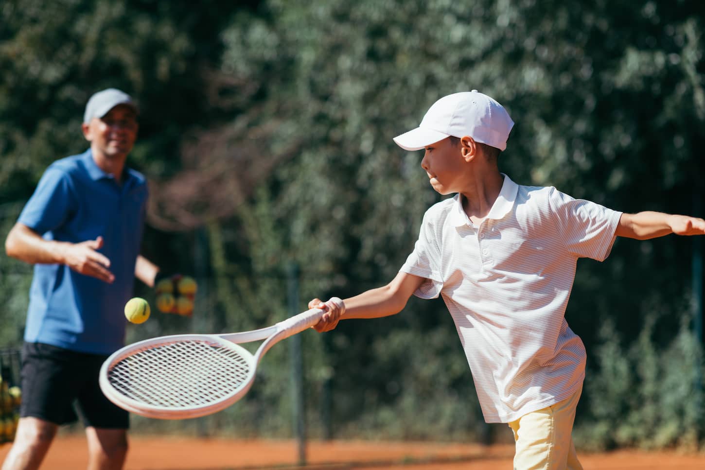 An image of a Tennis Instructor with a Boy Having a Tennis Lesson on a Clay Court.