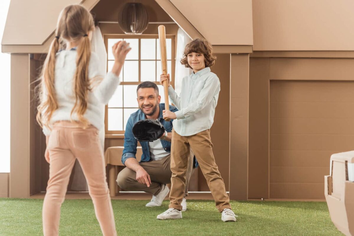 An image of a father teaching his kids how to play baseball in front of a cardboard house.