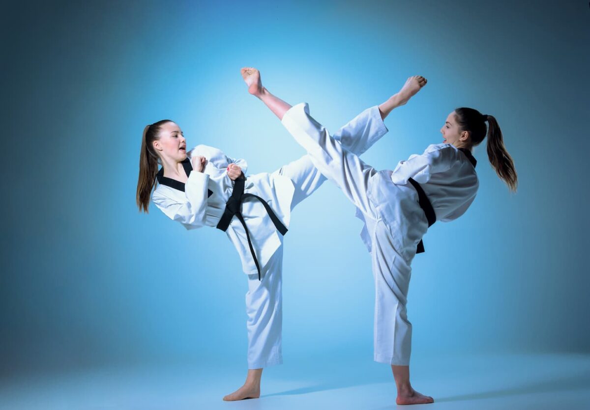 An image of a studio shot of two fit young women dressed in white kimono training in karate martial arts on a blue background.