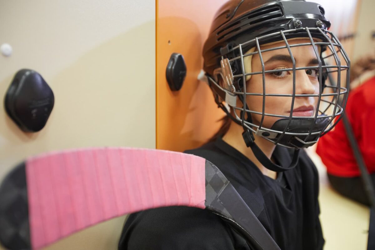An image of a beautiful young woman wearing hockey gear and looking at the camera while posing in the locker room.