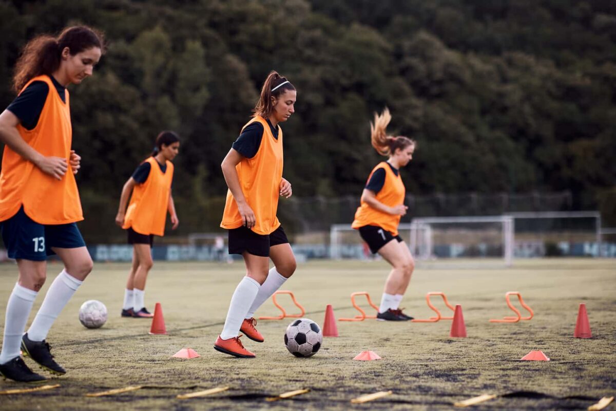 An image of a Female soccer player leading the ball among cones during sports training at the stadium.